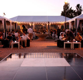 A photo of an outdoor party with a dance floor in the foreground and a large Made in the Shade tent in the background. A fountain is at the center of the photograph.